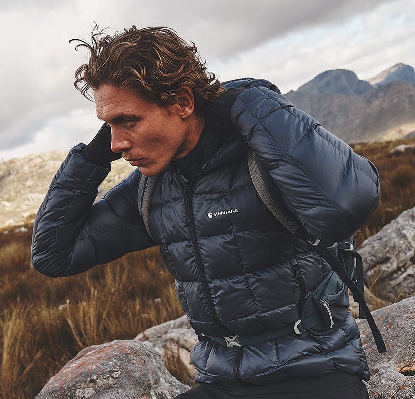 A man wearing a dark navy Montane insulated jacket sits on rocks in a mountainous outdoor setting, with his hand on his head in a contemplative pose.