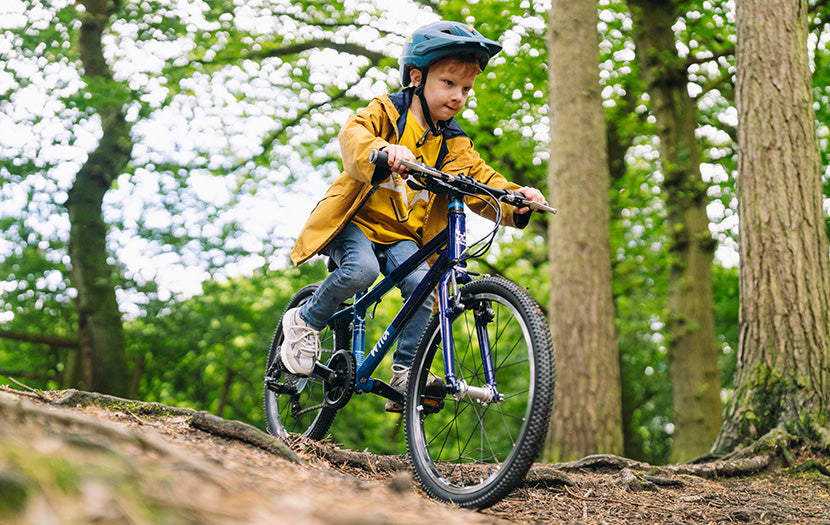 Child wearing a helmet and yellow jacket riding a blue mountain bike through a woodland trail.