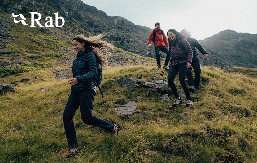Four people hiking up a rocky grassy mountain slope, with the Rab logo in the top-left corner. The foreground hiker wears a dark navy puffer jacket and dark pants, while others in red and purple jackets follow behind her on the trail.