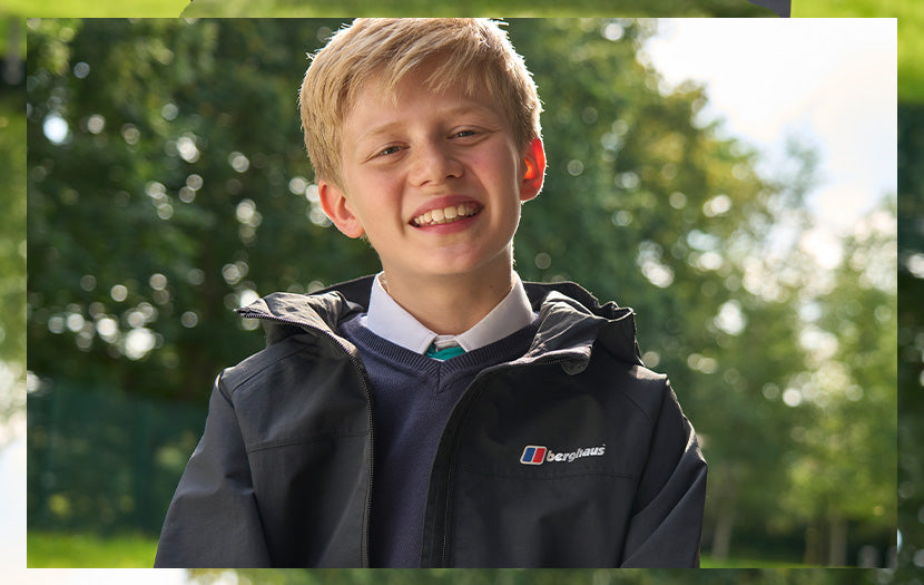 A smiling young boy wearing a black Berghaus windbreaker jacket over a navy sweater and white collared shirt, photographed outdoors with a blurred green background.