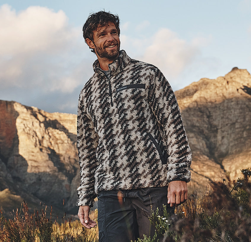 A man wearing a black and beige patterned fleece pullover jacket stands outdoors in a mountain landscape.