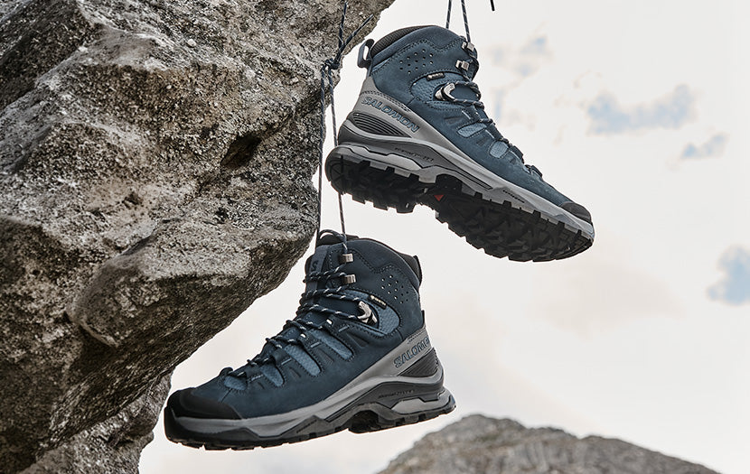 A pair of dark slate blue and gray Salomon mid-height hiking boots hanging from ropes against a large rock face with a cloudy sky in the background.